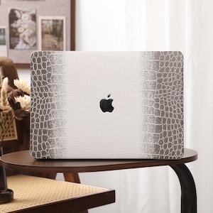 Puede incluir: Una funda blanca para portátil con un estampado de piel de cocodrilo en tonos grises. El logotipo de Apple es visible en negro. La funda está sobre una mesa marrón oscuro.