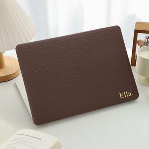 May include: A dark brown laptop case with a textured crocodile skin pattern and the name Ella. printed in gold lettering on the bottom right corner. The case is designed to fit over the exterior lid of a laptop, providing a protective cover.