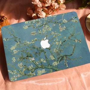 May include: A blue laptop case with a painting of a blossoming almond tree branch. The case has a white Apple logo. The artwork is in the style of Vincent van Gogh. A cup of coffee and a decorative gold frame are in the background.