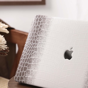 May include: A white laptop case with a faux crocodile skin pattern. The case has a gradient effect, transitioning from white to gray. The Apple logo is visible on the case. The laptop is resting on a wooden surface.