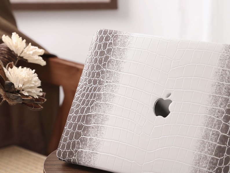 May include: A white laptop case with a faux crocodile skin pattern. The case has a gradient effect, transitioning from white to gray. The Apple logo is visible on the case. The laptop is resting on a wooden surface.