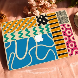 May include: A colorful laptop case with a patchwork design. The case features various geometric patterns in shades of blue, orange, black, pink, and green. The Apple logo is visible. A cup of coffee sits on a saucer nearby.