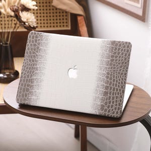 May include: A laptop computer with a white and brown crocodile-textured case, featuring the Apple logo. The laptop is on a small, round wooden table. The case has a central white panel with brown textured sides.