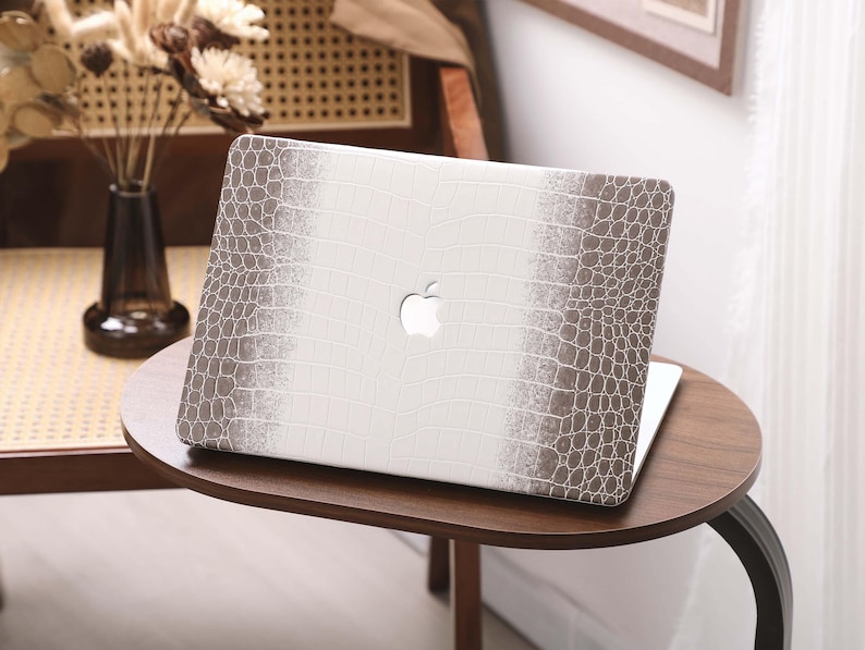 May include: A laptop computer with a white and brown crocodile-textured case, featuring the Apple logo. The laptop is on a small, round wooden table. The case has a central white panel with brown textured sides.