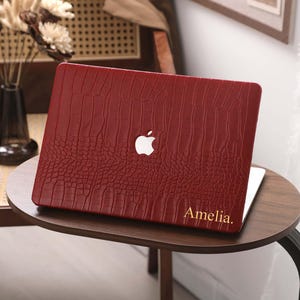 Puede incluir: Una funda para portátil de color burdeos con un patrón texturizado similar a la piel de cocodrilo. El logotipo de Apple es visible, junto con el nombre "Amelia" en letras doradas. La funda está sobre una pequeña mesa redonda de madera.