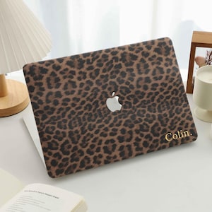 Op de afbeelding: Een laptop met een bruin en zwart luipaardprint hoesje. Het Apple-logo is zichtbaar. De naam "Colin" is in goud gedrukt in de rechter benedenhoek. De laptop ligt op een wit oppervlak.