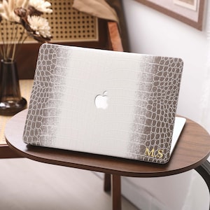 May include: A white and brown laptop case with a crocodile skin pattern. The case has a white center with brown edges and the Apple logo. Gold initials "M.S" are on the bottom right corner. The laptop is on a small, round, wooden table.
