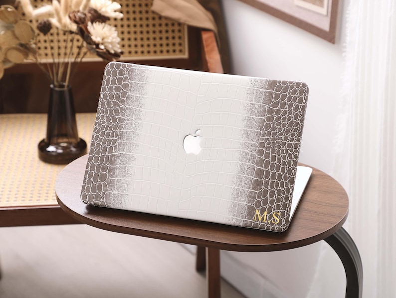 May include: A white and brown laptop case with a crocodile skin pattern. The case has a white center with brown edges and the Apple logo. Gold initials "M.S" are on the bottom right corner. The laptop is on a small, round, wooden table.