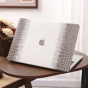 May include: A white laptop with a gray crocodile-textured design on the cover, featuring the Apple logo. The laptop is placed on a dark brown, round side table. The design has a gradient effect.