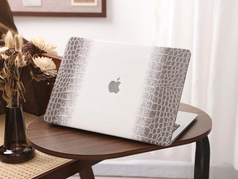 May include: A white laptop with a gray crocodile-textured design on the cover, featuring the Apple logo. The laptop is placed on a dark brown, round side table. The design has a gradient effect.