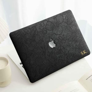 Puede incluir: Funda negra para portátil con diseño floral en relieve. La funda tiene un recorte para el logotipo de Apple y las iniciales doradas "SK.". El portátil está abierto sobre una superficie blanca con una vela y una lámpara.