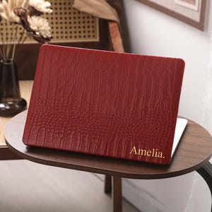 Può includere: Una custodia per computer portatile rosso scuro con una texture a pelle di coccodrillo. La superficie della custodia presenta il nome Amelia impresso con un carattere serif color oro, posizionato nell'angolo in basso a destra della cover.