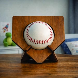 Könnte beinhalten: Ein Baseball, der in einem holzfarbenen Rahmen in Form einer Homeplate präsentiert wird. Der Baseball ist weiß mit roten Nähten. Der Rahmen besteht aus dunkelbraunem Holz und hat einen Ständer, um ihn aufrecht zu halten. Der Hintergrund ist unscharf.