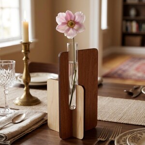 May include: A wooden flower vase with a clear glass tube holding a pink flower. The vase has a dark brown back and a lighter wood front. The vase is on a wooden table with a candle and silverware.