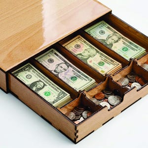 Puede incluir: Un cajón de caja registradora de madera con compartimentos para billetes y monedas. El cajón está abierto, revelando pilas de billetes de uno, cinco, diez y veinte dólares, junto con una colección de monedas. La madera tiene un acabado marrón claro.