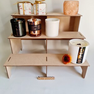 May include: A wooden tiered display stand with four shelves, featuring a variety of candles, gift boxes, and a salt lamp. The stand is made of natural wood and has a simple, rustic design. The candles are in various sizes and colors, including black, white, and rose gold. The gift boxes are wrapped in white paper with floral designs. The salt lamp is a natural pink color and is shaped like a rock. The stand is labeled "Laser Wood Lab".