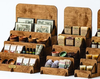 May include: Wooden tiered display stands showcasing various artisanal products. The stands hold framed prints, small wooden bowls, packaged goods, essential oils, soaps, candles, and jars of honey. The displays are made of light brown wood.