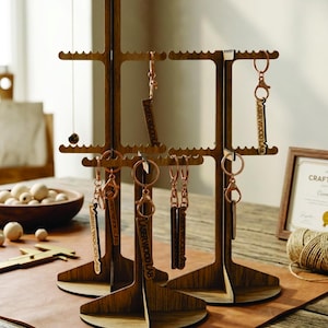 May include: Wooden key and jewelry display stands with multiple levels. The stands hold keychains and a necklace. The keychains are engraved with the text "LASER WOOD LAB". The stands are on a brown leather surface.