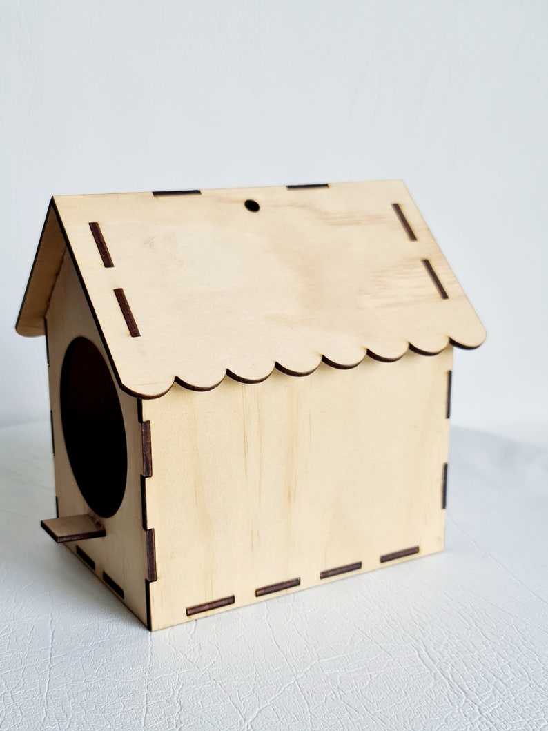 May include: A wooden birdhouse with a scalloped roof and a round entrance hole. The birdhouse is made of light brown wood and has a small perch on the front.