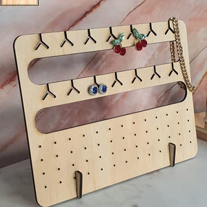 May include: Wooden earring display stand with two rows of hooks and a bottom row of small holes. The stand has two small feet and is made of light wood. The stand is decorated with red cherry earrings, blue earrings, and a gold chain.