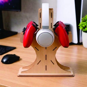 以下が含まれることがあります： 木製のゲーム用ヘッドセットとコントローラーのスタンド。スタンドには、白とグレーのヘッドセットと2つの赤いコントローラーが収納されています。スタンドは明るい色の木材で作られており、木の形をしています。黒いマウスが机の上にあります。