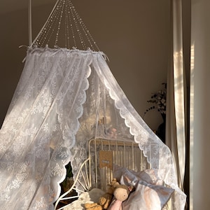 May include: A white lace canopy hangs above a white metal bed. The bed is made with pillows, a doll, and a teddy bear. An open book is on the bed. The room is lit by sunlight.