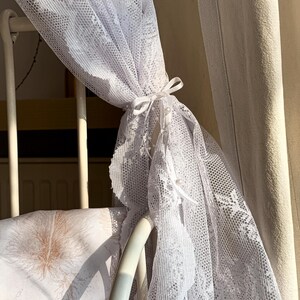 May include: White lace canopy draped over a white metal cot with a brown and white patterned pillow. The canopy is tied with a white ribbon.