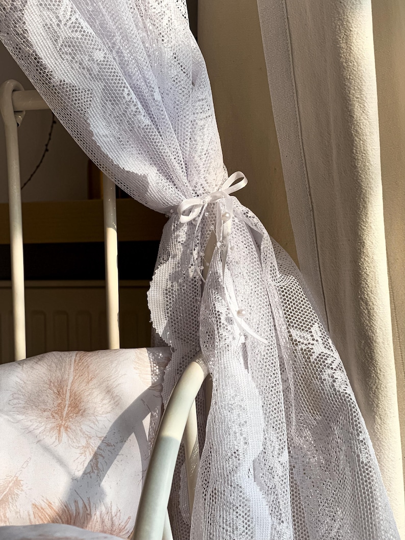 May include: White lace canopy draped over a white metal cot with a brown and white patterned pillow. The canopy is tied with a white ribbon.