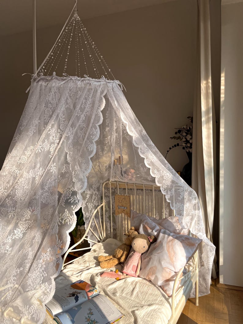 May include: White lace canopy hanging over a white bed frame with a white and pink patterned pillow and two stuffed toys. The bed is decorated with a book and a sign that says "Dreamer".