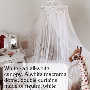 May include: White macrame canopy with a white dome and double curtains made of neutral white viscose and snow-white mesh. The canopy is hanging over a white bed with a stuffed giraffe toy.