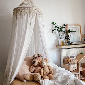 May include: A white canopy bed with a white crocheted top and a brown bedspread. Two teddy bears are sitting on the bed, one wearing a flower crown.