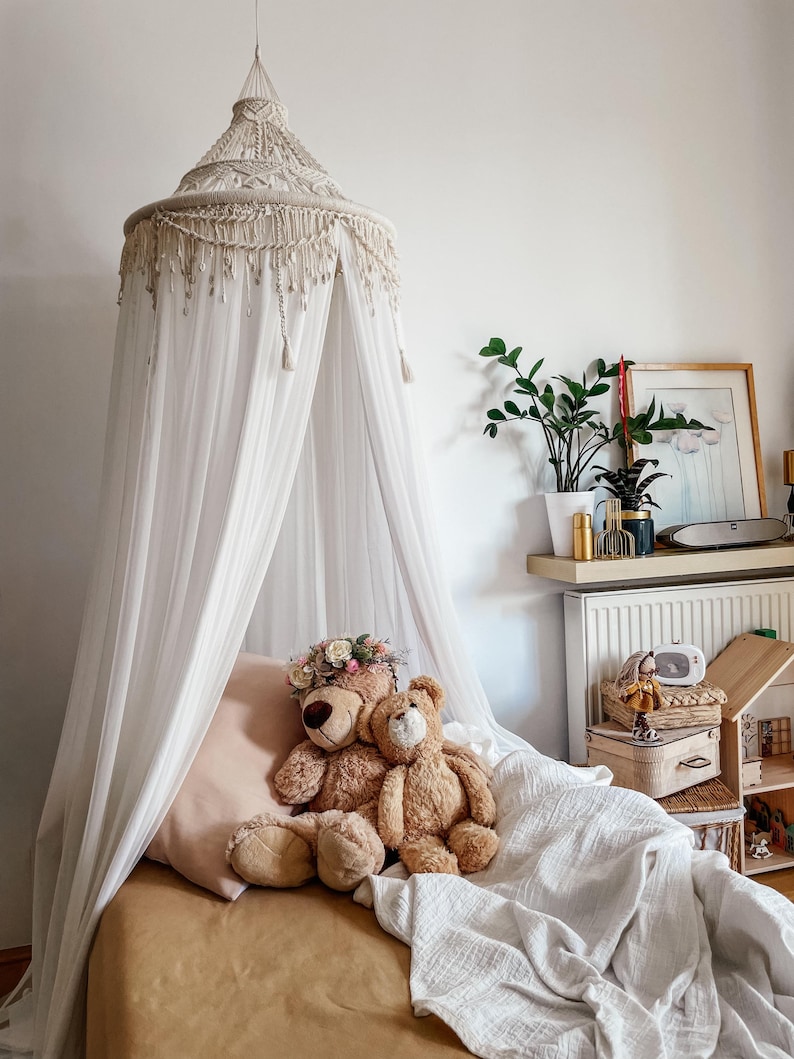 May include: A white canopy bed with a white crocheted top and a brown bedspread. Two teddy bears are sitting on the bed, one wearing a flower crown.