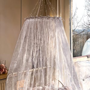 May include: White lace canopy with a beaded top, hanging over a white cot with a white lace blanket and pillows. The canopy is made of delicate lace and has a romantic feel.