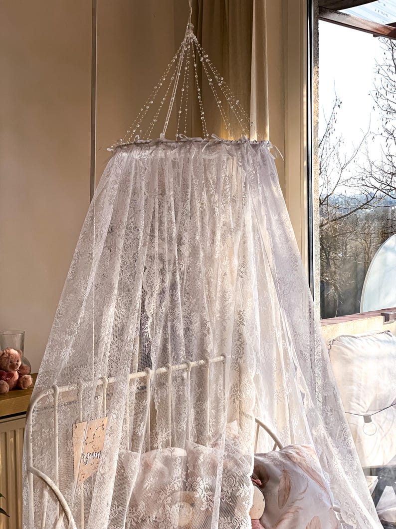 May include: White lace canopy with a beaded top, hanging over a white cot with a white lace blanket and pillows. The canopy is made of delicate lace and has a romantic feel.