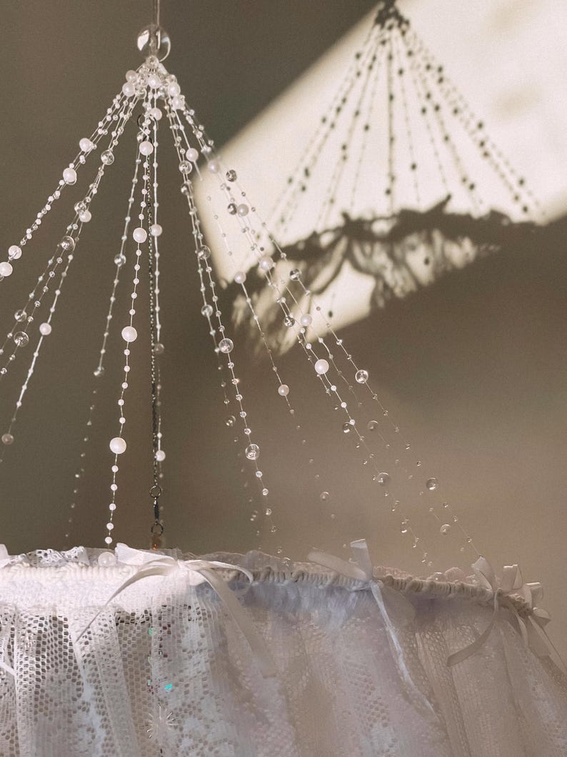 May include: A white lace canopy with a beaded chandelier hanging from the centre. The beads are clear and reflect light.