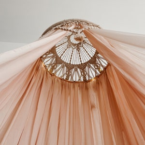 May include: A light pink canopy with a macrame top. The canopy is made of a sheer fabric and is hung from the ceiling.