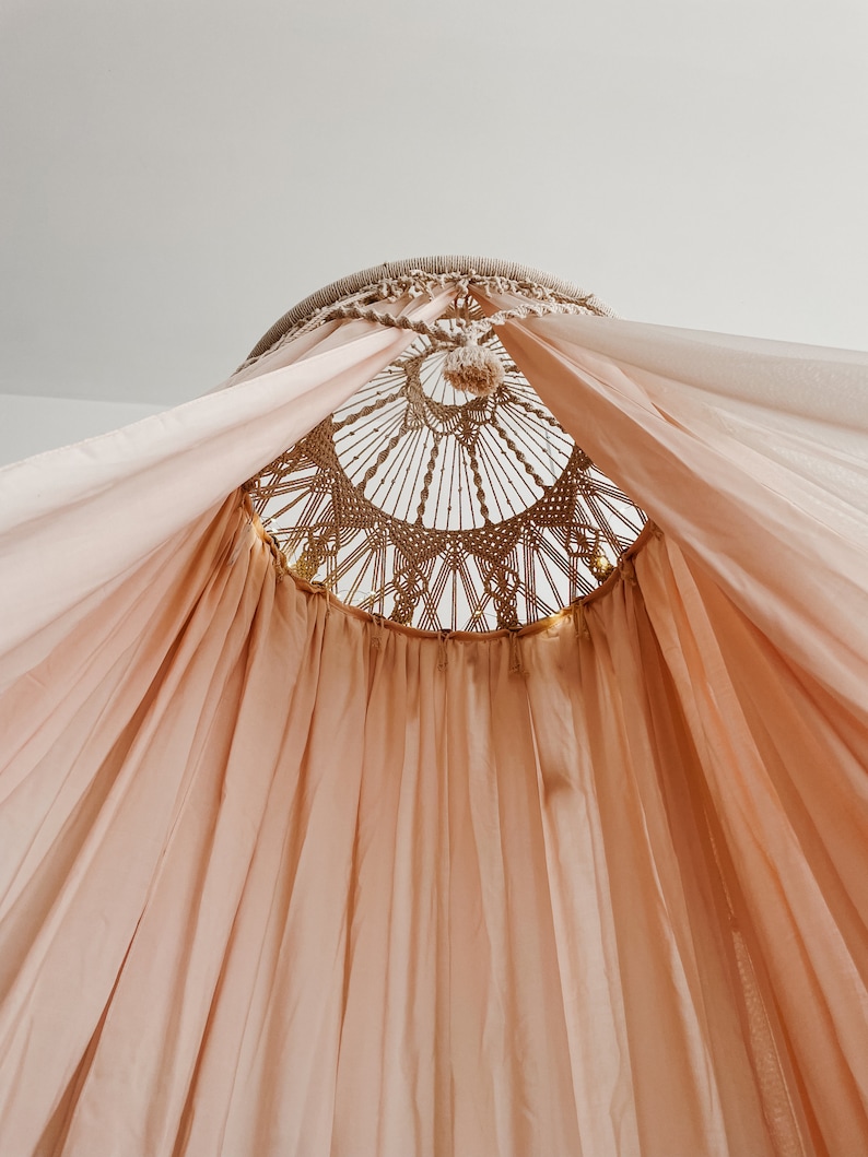 May include: A light pink canopy with a macrame top. The canopy is made of a sheer fabric and is hung from the ceiling.