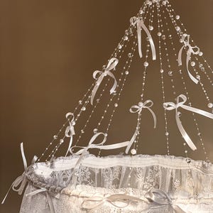 May include: White lace canopy with a crystal and ribbon mobile hanging from the top. The mobile features white ribbons tied in bows and clear crystal beads.