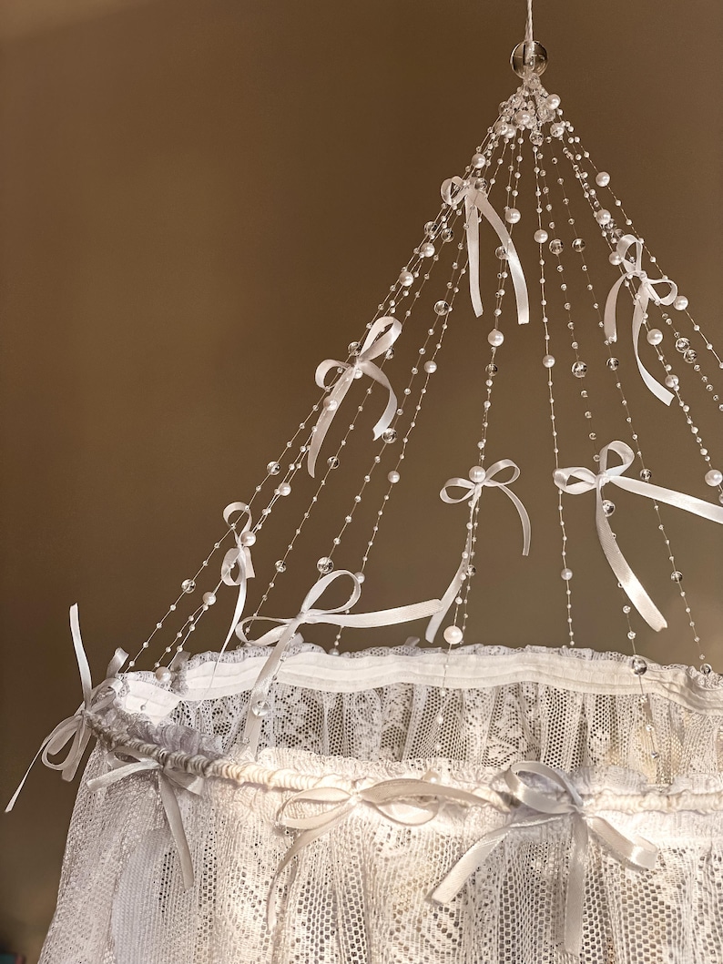 May include: White lace canopy with a crystal and ribbon mobile hanging from the top. The mobile features white ribbons tied in bows and clear crystal beads.