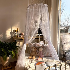 May include: A white lace canopy hangs over a white metal bed frame with a white and pink patterned pillow. Two stuffed toys, a teddy bear and a doll, are on the bed. A book is open on the bed.