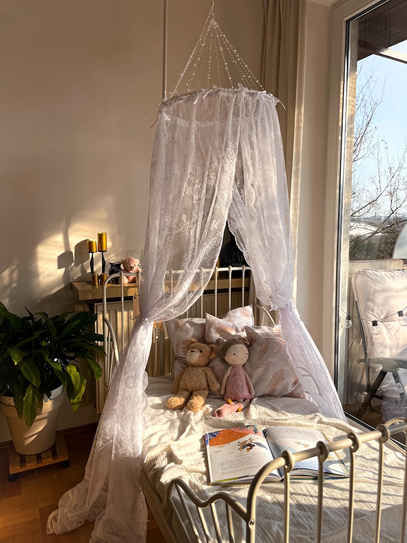 May include: A white lace canopy hangs over a white metal bed frame with a white and pink patterned pillow. Two stuffed toys, a teddy bear and a doll, are on the bed. A book is open on the bed.