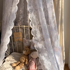 May include: A white lace canopy draped over a bed with a white metal frame. Two stuffed toys, a brown teddy bear and a pink doll, are lying on the bed. The canopy has a scalloped edge and a floral pattern. The bed is covered in a white blanket.
