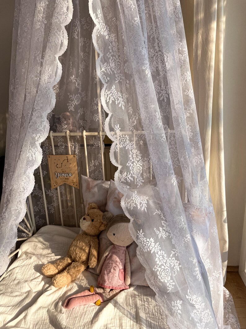 May include: A white lace canopy draped over a bed with a white metal frame. Two stuffed toys, a brown teddy bear and a pink doll, are lying on the bed. The canopy has a scalloped edge and a floral pattern. The bed is covered in a white blanket.