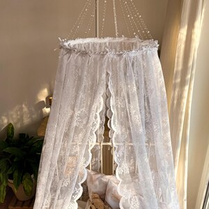 May include: White lace canopy with a scalloped edge, hanging over a cot with a stuffed animal and doll inside. The canopy is decorated with a string of white beads.