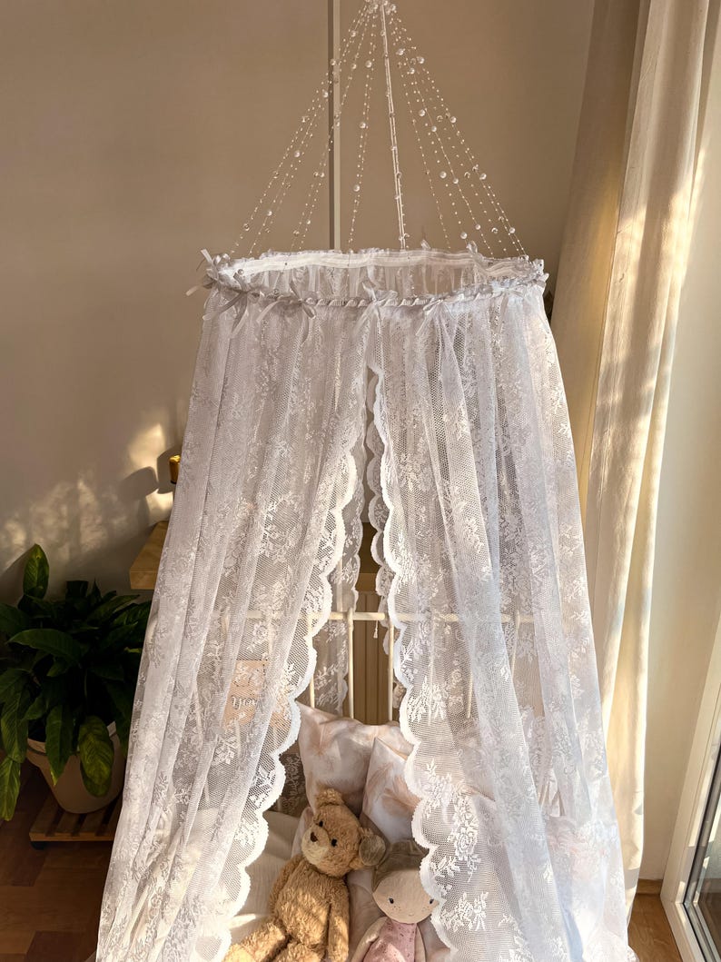 May include: White lace canopy with a scalloped edge, hanging over a cot with a stuffed animal and doll inside. The canopy is decorated with a string of white beads.