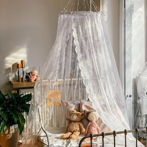 Puede incluir: Un dosel de encaje blanco cuelga sobre una estructura de cama de metal blanco con una almohada estampada en blanco y rosa, un oso de peluche marrón y una muñeca rosa. La cama está decorada con un libro y un cartel que dice "Eres amado".