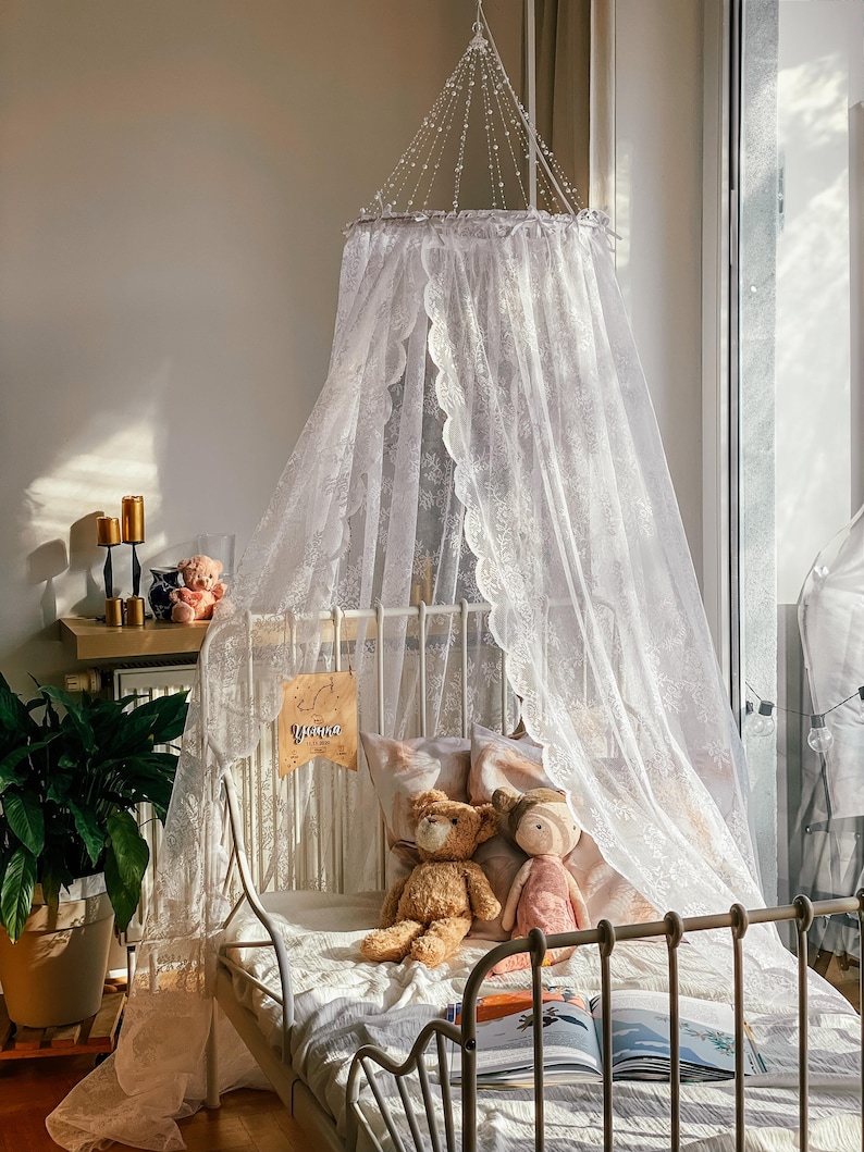 May include: A white lace canopy hangs over a white metal bed frame with a white and pink patterned pillow, a brown teddy bear, and a pink doll. The bed is decorated with a book and a sign that says "You are loved".