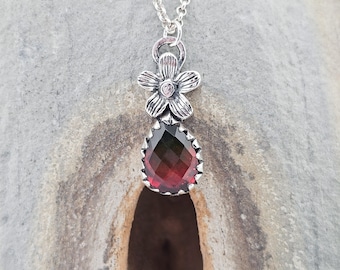 May include: A silver necklace featuring a flower pendant with a small gemstone at its center. Below, a teardrop-shaped, faceted gemstone displays a gradient of red and green hues. The necklace is displayed against a textured gray background.