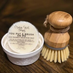 May include: A white container with a round, off-white soap labeled "Crazy Girl Soaps" and "YOU PUT A BRUSH ON ME." A wooden dish brush with light-colored bristles is next to the soap. The soap is eco-friendly.