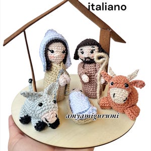 Può includere: Un presepe all'uncinetto con Maria, Giuseppe, Gesù bambino, un asino e un bue, su una base di legno. Le figure sono beige, marroni, blu e grigie. Il testo "Pattern Pdf italiano" è visibile.